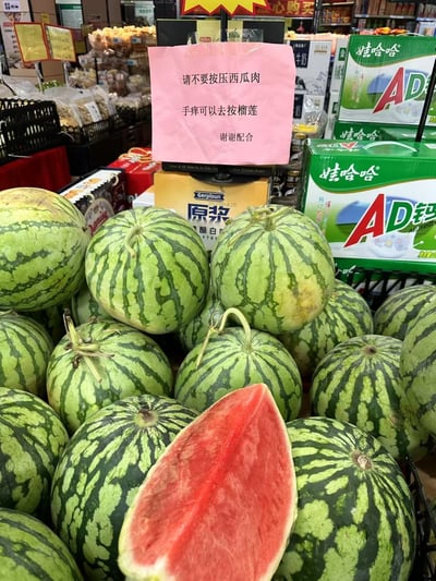 这是一张水果店或超市内的实拍图，展示了西瓜售卖区放置的一个幽默提示牌。告示上写着'请不要按压西瓜肉，手痒可以去按榴莲，谢谢配合'，劝阻顾客不要用手捏西瓜，巧妙调侃'手痒'的人可以去按坚硬的榴莲来解解痒，语气轻松风趣，表达了商家的善意无奈。由双关和反差制造幽默，适合网上传播和讨论。