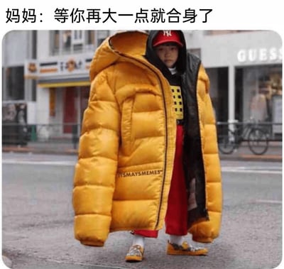 这张图片展示了一个站在城市街道上的小孩，他穿着一件尺寸严重偏大的黄色连帽羽绒服，羽绒服的衣长和袖长都远远超过小孩的身材，袖子拖在地面上，小孩仅露出头部、下半身和鞋子，脸上露出无奈又委屈的神情。图片上方配有文字“妈妈：等你再大一点就合身了”，精准戳中了很多家长给孩子买衣服时，为了让孩子能多穿几年而刻意买大好几号的普遍生活场景，用夸张的视觉效果放大了这种日常行为的搞笑感，引发观者的共鸣。背景是普通的城市街道，能看到路边的商铺和自行车，整体氛围充满了生活化的趣味。