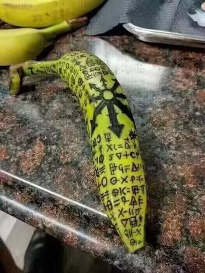 这张图展示了一根放置在大理石纹理台面上的黄绿色香蕉，香蕉表皮被用黑色颜料精心绘制了大量类似神秘符文、未知字符的内容，中间位置还有一个由对称箭头组成的星形特殊符号，整体风格充满神秘、猎奇感，仿佛是虚构作品里的仪式道具。背景中还能看到两根普通的黄色香蕉以及一个金属容器，与这根被涂鸦的香蕉形成对比，凸显其特殊性。图中没有双关、谐音梗或暗示类内容。