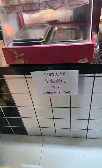 图片展示了一家餐饮店的内部场景，主要聚焦于一个红色的食品保温展示柜下方。展示柜看起来是空的，下方的黑白格子瓷砖墙上贴着一张手写的招聘告示，内容为“招聘女同学包馄饨 电话”，其中电话号码部分被模糊处理。