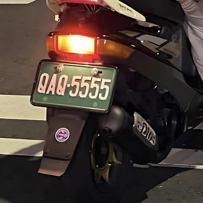 图片特写摩托车尾部，绿色车牌上显示“QAQ-55555”，尾灯亮着红黄相间的灯光。其中“QAQ”是网络流行的哭泣表情符号，而“55555”在中文网络语境中因谐音“呜呜呜”也常用来表示哭泣，两者结合让车牌看起来像在“哭”，形成幽默有趣的视觉效果。