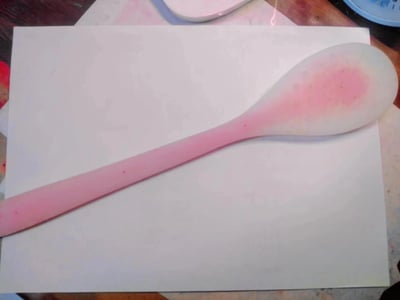 图片中展示了一张白色的纸张平铺在木质桌面上，纸张中央放置着一把勺子。勺子的手柄部分为粉红色，勺头部分则是白色中带有一块明显的粉红色污渍，整体呈现出柔和的色彩对比。背景中隐约可见其他物品的边缘，但焦点集中在勺子和白纸上，构图简洁清晰，主要展现了这一日常物品的静态状态。