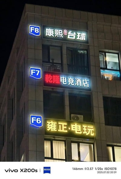 这是一张夜晚拍摄的商业楼外立面照片，展示了三层商家的发光招牌：F6楼层是「雍正·电玩」，暖黄色发光字体；F7楼层是「乾隆电竞酒店」，红白色发光字体；F8楼层是「康熙台球」，白绿色发光字体。三个商家分别以清朝三位知名帝王的名字命名，形成了连贯又有趣的系列感，制造出帝王“跨界”经营现代休闲娱乐生意的反差萌，十分吸睛。图片底部带有vivo X200s的拍摄水印，标注了拍摄参数与时间：2025年7月2日20:39。