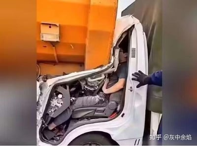 这是一张利用特殊拍摄角度形成的视觉错位照片：画面中一辆白色小型车辆的驾驶室被旁边的大型橙色车辆挤压变形，从当前视角看去，车内司机的身体与车辆破损后露出的内部零件恰好重合，营造出司机身体和车辆结构“融为一体”的错觉。画面右侧还有一只手伸向驾驶位，看似是要帮助司机，实则是拍摄角度带来的趣味视觉效果，并非真实的身体与车辆融合，场景看起来惊险却充满了错位的趣味性。