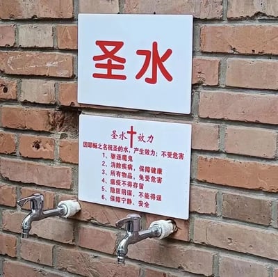 图片展示了一面红砖墙上挂着的两块白色标语牌。上方标语牌用红色大字写着“圣水”，下方标语牌标题为“圣水+效力”并带有十字架符号，内容详细列出了六项所谓“效力”：驱逐魔鬼、消除疾病保障健康、所有物品免受危害、瘟疫不得存留、隐匿阴谋不能得逞、保障宁静安全。标语牌下方安装有两个银色水龙头，整体给人一种将普通自来水赋予宗教神秘色彩的感觉。图片中无明显双关、谐音梗或暗示内容，主要呈现带有宗教元素的民间信仰标语。