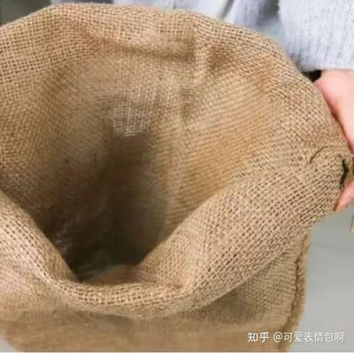 图片为特写视角，展示一只手（穿着浅灰色毛衣）正握着一个棕色粗麻布袋的开口处，袋口被撑开，内部呈深色，麻布袋的编织纹理清晰可见，整体画面简洁，突出日常物品的质朴质感。