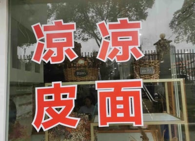 这是一张线下小吃店玻璃门的实拍静态图，玻璃外侧贴有红底白边的醒目大字，以两行排版呈现，上行是两个“凉”字，下行是“皮”“面”字，组合起来明确告知店铺主营“凉皮”与“凉面”两款凉食。玻璃表面反射出店外的树木、金属围栏和城市建筑，玻璃内侧可以看到两个带有英文标识的编织装饰，分别标注“Plants”和“HOME”，还能看到一个模糊的店内人员身影，整体场景是日常的街头小吃店门面，风格朴素接地气，用于直观展示店铺的主营品类。