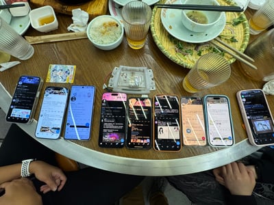图片展示了广州南村万博一场面基聚会的餐桌场景。圆形餐桌上整齐摆放着8-9部智能手机，屏幕亮着显示不同应用界面，周围散落着米饭碗、剩菜盘、玻璃杯（内有黄色饮品）、筷子、纸巾等餐具和食物，桌边可见几人的手和手臂，整体呈现出热闹的线下聚会氛围。