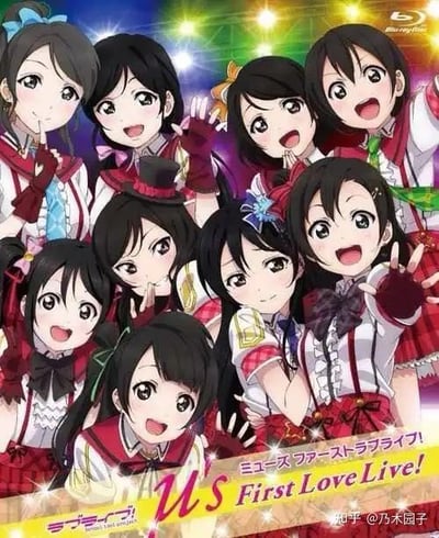 图片为《Love Live! School Idol Project》中校园偶像团体μ's的9名成员官方Blu-ray封面。成员们身着红白为主色调的华丽演出服，佩戴蝴蝶结、礼帽等装饰，面带灿烂笑容摆出多样活泼姿势（如挥手、比耶、比心）。背景是渐变紫色与彩色光斑交织的舞台效果，营造出演唱会的热烈氛围。封面底部有标题文字“μ's First Love Live!”及日文名称，右上角可见Blu-ray Disc标志。