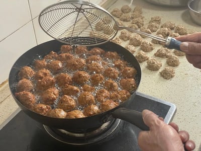 这张图片展示了一个家庭厨房的烹饪场景，有人正在油炸肉丸子。锅中装满了正在翻滚的金黄色肉丸子，旁边的台面上摆放着许多待炸的生肉丸子。一只手正拿着金属漏勺准备翻动锅中的丸子，整个画面充满了生活气息和美食的诱惑。