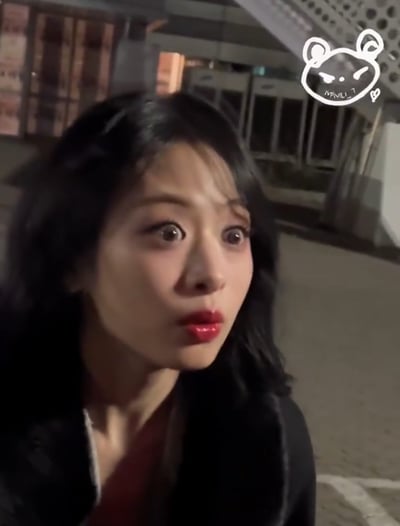 这张图片是在夜晚户外拍摄的，焦点是一名有黑长发、穿冬天外套的女性。画面整体光线较暗，背景为街头公共空间，右上角贴有卡通手绘熊头表情贴纸元素，增加了整体的可爱和俏皮氛围，给人青春和治愈的感觉。适合用作朋友圈、生活分享类图片。