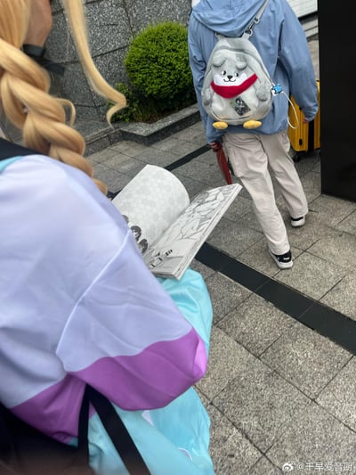 这是一张户外实拍的日常照片，前景是一位cos《LoveLive!Superstar!!》千早爱音的爱好者，有着金色麻花辫，穿着紫白拼色的cos服装，正低头翻看一本二次元线稿画集；背景里有一位背着可爱灰色动物造型毛绒背包（背包上还挂着二次元角色挂饰）的人，手持红色雨伞，拉着黄色行李箱，整体场景充满二次元氛围，看起来像是漫展场外的相关日常。