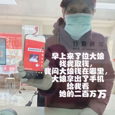 图片中一位戴着浅蓝色口罩和红色帽子的大娘，身穿深色外套，正手持一部智能手机面向镜头。手机屏幕显示红色背景界面，上方有白色文字“2000000万”及一个人物头像图标。图片右侧配有文字说明场景：“早上来了位大娘找我取钱，我问大娘钱在哪里，大娘拿出了手机给我看她的二百万万”。场景疑似发生在银行或类似服务场所，背景可见室内环境及部分标识。整体通过真实人物与手机屏幕内容的结合，呈现出因金额单位误解产生的生活趣事，带有幽默诙谐的氛围。