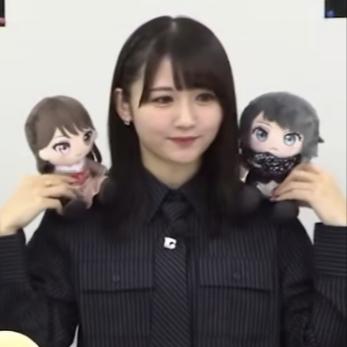 图中是一位留着齐刘海的黑发女偶像，身穿深色细条纹衬衫搭配领带，面带温柔微笑，双手分别举着两个Q版卡通毛绒玩偶：一个是双马尾造型的软萌女孩玩偶，另一个是戴着面罩的酷感深色头发玩偶。整体画面温馨可爱，场景看起来像是在直播或线下活动中向观众展示相关周边产品。