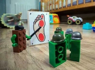 画面中，一个棕色身体、黑色帽子的“老乐高”正手持黑色教鞭，站在一块画有红色脚印图案（脚印上有两个红色圆点标记）的白板前教学。白板前整齐站立着四个绿色身体、灰色头盔的“小乐高士兵”，它们正专注地抬头听讲。背景中散落着恐龙玩具、彩色积木、玩具车等物品，营造出充满童趣的玩具场景。图中通过拟人化手法，将乐高玩具塑造成“教学埋伏人类”的趣味画面，红色脚印上的圆点可能暗示人类脚部的“弱点”或“埋伏目标”，充满幽默创意。