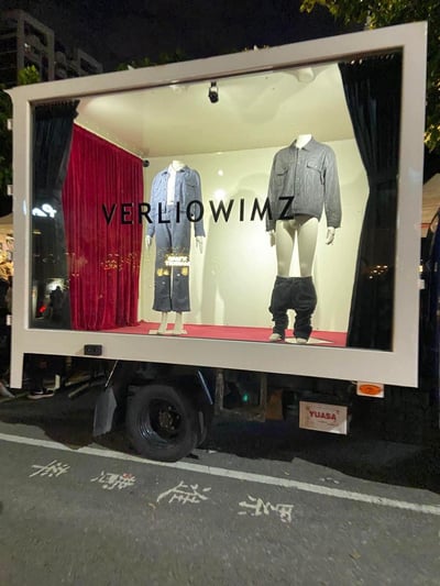 夜晚场景下的移动时装展示车，展示窗内有两个人体模型穿着时尚牛仔服装。左侧模型穿着蓝色牛仔套装，右侧模型穿着灰色牛仔夹克和黑色蓬松裤装。展示背景采用红色幕布和白色墙面的组合，顶部有射灯照明。展示车外部可以看到城市夜景的模糊灯光，底部有YUASA的标识。