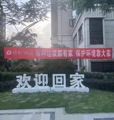 这是一张拍摄于保利物业管辖小区内的实景照片，前景的草坪上放置着白色的立体字标识“欢迎回家”，后方的红色横幅上印有保利物业的品牌标识以及垃圾分类宣传标语“每种垃圾都有家 保护环境靠大家”。背景可见小区的高层住宅楼和绿植树木，整体场景温馨正式，是社区常见的宣传布置，图中没有双关、谐音梗等特殊内容。