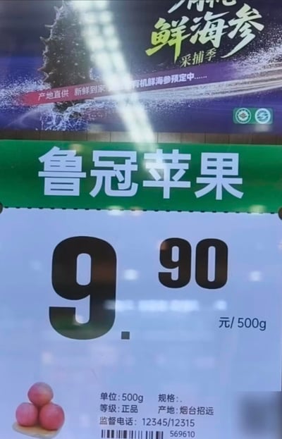 图片展示了一个超市或水果店的商品价格牌，主要内容为绿色背景的"鲁冠苹果"价格标签，显示价格为9.90元/500g，下方标注了单位、等级（正品）、产地（烟台招远）及监督电话。标签上方有一个部分可见的紫色背景广告，内容为"鲜海参 采捕季"，并提到"产地直供 新鲜到家"等字样。