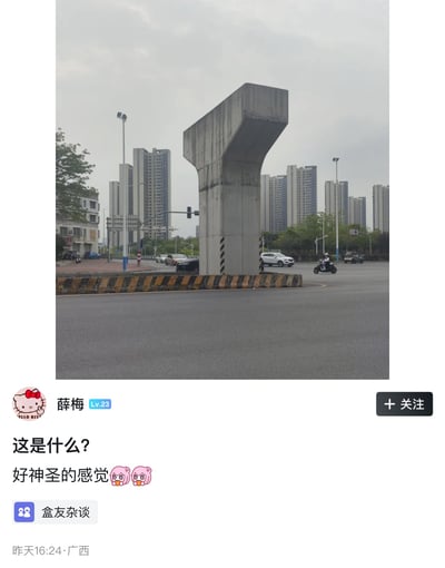 这张图是社交平台截图，上半部分为实拍画面：城市道路路口的中央立着一个大型灰色混凝土桥墩，上宽下窄造型方正，底部周围环绕着黄黑相间的警示路障，周边道路上有行驶的汽车、电动车，背景是成片的高层居民住宅楼与路灯，天空阴沉。下半部分是小黑盒平台的用户发帖内容，头像是Hello Kitty的用户“薛梅”（等级Lv.23）配文提问“这是什么？好神圣的感觉”，附带两个惊讶的粉色表情，帖子关联话题为“盒友杂谈”，发布地址为广西，发布时间为昨日16:24。该混凝土墩因外形与游戏《我的世界》中铁傀儡的身体部分高度相似，被网友玩梗调侃为铁傀儡的半成品，只要在顶部放置南瓜头外壳，外层水泥就会脱落变成铁傀儡，可以在夜晚怪物多的时候应急使用。