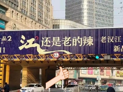 这是一张经过后期加工的城市街景实拍图，画面主体是跨路桥上的户外广告：深蓝色背景上印着醒目的金色「江」字，搭配白色文字「还是老的辣」，右侧标注「老汉江 新品」，左侧有「品2」「11」字样。广告巧妙运用谐音梗，将俗语「姜还是老的辣」中的「姜」替换为「江」，结合「老汉江」的品牌名，形成趣味营销效果。图片后期添加了一个卡通点赞手势，突出对这个谐音梗的认可。背景是繁华的城市商业街，有高楼建筑、行驶的车辆、行人，交通信号灯显示绿色，桥洞下的限高标识为4.4m。