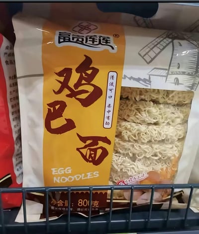 图片展示了超市货架上的一袋方便面产品，品牌为"富贵莲"，产品名称为"鸡巴面"。包装主体为黄色，左侧有醒目的红色"鸡巴面"大字，右侧为透明窗口展示面条实物。包装上还有"清淡可口 柔中有劲"的产品描述和英文翻译"EGG NOODLES"。净含量为800克。

图片存在明显的低俗名称问题，"鸡巴"在中文中是粗俗俚语，用作食品名称涉嫌违反公序良俗。