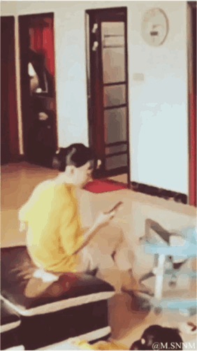这是一个制造视觉错觉的动态GIF，画面中一位身穿黄色上衣的女生坐在室内的带垫凳子上，低头专注看着手机。由于凳子黑色坐垫的位置和形态，视觉上看起来像是女生的裤子滑落下来，形成了搞笑的反差效果，实际只是凳子的垫子，场景为普通的室内客厅环境。