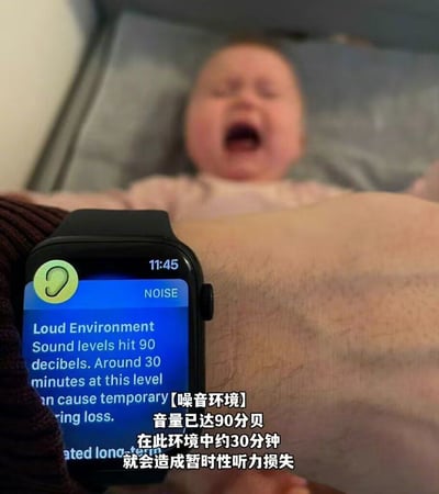 图片展示了一个有趣的生活场景：前景是一只智能手表，屏幕上显示着噪音警报，提示环境声音已达到90分贝，长时间暴露可能导致暂时性听力损失。背景中可以看到一个婴儿躺在床上大哭，显然是婴儿的哭声触发了这个噪音警报。这种将现代科技产品与婴儿日常结合的画面，形成了一种幽默又略带无奈的对比，生动展现了育儿生活中的小插曲。