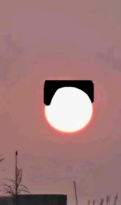 这是一张经过创意修改的落日照片，原本散发着暖红色光晕的圆形落日，被添加了一块黑色的齐刘海形状图案，瞬间让太阳拥有了拟人化的可爱造型，仿佛留着齐刘海的卡通形象。背景是粉紫色调的黄昏天空，下方能看到植物和建筑的边缘，整体画面充满了搞笑又治愈的趣味感，给普通的日落场景增添了十足的创意。