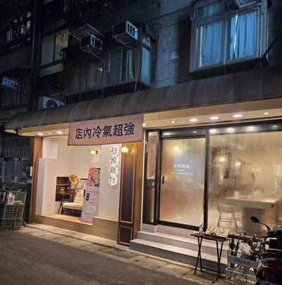 这是一张夜晚街头的店铺实拍图，展示了位于居民楼底层的“初殿锅物”火锅店。店铺外观简约温馨，暖黄色灯光从店内透出，营造出舒适的氛围。门口悬挂着显眼的“店内冷气超强”横幅，用直白的宣传点吸引食客；门柱、玻璃门、店外宣传立牌、小桌标识以及停放的摩托车上都印有“初殿锅物”的店名。背景是带有外挂空调机的居民楼，整体呈现出夜晚城市里接地气的街边小店景象，既突出了火锅店的属性，又强调了店内凉爽的环境，适合想要吃暖锅同时享受舒适温度的顾客。
