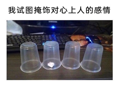 这是一张图文结合的趣味梗图，上方配有文字“我试图掩饰对心上人的感情”，下方展示了四个倒扣在深色桌面的透明塑料杯，其中从左往右数第二个杯子内部藏有一个白色的爱心图案，背景是带有键盘和鼠标的办公桌面场景。整幅图用杯子掩饰爱心的具象画面，生动又幽默地比喻了暗恋者想要隐藏对心上人的喜欢，却还是不经意间露出破绽的状态，精准戳中了暗恋时那种羞涩、想藏又藏不住的细腻情绪。