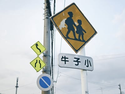 这张图呈现了户外道路旁的交通标识场景，右侧白色立柱上方是带有磨损痕迹的黄底黑边注意儿童通行警示标识，标识下方的白色矩形路牌被倒置，显示文字为“牛子小”；左侧灰色电线杆上从上到下依次设有向左急转弯警示标识、车道收拢变窄警示标识、蓝底白边的禁止长时间停车禁令标识，背景是阴天的浅灰色天空，还能看到通信基站、电线杆与电线。其中“牛子”是中国部分地区方言中对男性生殖器的俗称，倒置路牌呈现出的这三个字属于低俗趣味网络梗，该路牌内容大概率为网友后期P图修改而成。