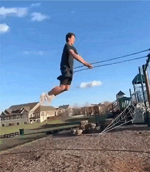 A humorous yet cringe-worthy sequence showing a person attempting an ambitious jump off a playground swing. The individual tries to leap over a rock formation but fails spectacularly, landing face-first in the gravel. They then slowly recover, getting on their hands and knees before crawling away from the scene, clearly embarrassed by the mishap. The suburban neighborhood setting with houses in the background adds to the everyday nature of the failed stunt.