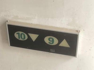 这是一张安装在墙面上的楼层指示牌实拍图，指示牌为黑底，带有夜光效果的标识：左侧是圆形框内的绿色数字“10”，搭配向下的三角箭头，代表前往10层的方向为向下；右侧是圆形框内的绿色数字“9”，搭配向上的三角箭头，代表前往9层的方向为向上。指示牌右下角还有“充电”“主电”“故障”三个状态标识，墙面有轻微的裂纹和掉漆痕迹。