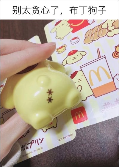 这是一张结合实拍与配文的趣味图，图片顶部配有黑色文字「别太贪心了，布丁狗子」。画面中一只手握着黄色的布丁狗公仔，将公仔的臀部对准下方的麦当劳和三丽鸥布丁狗联名贴纸，公仔臀部印有两个星号图案来模拟臀部形态，搭配配文制造出调侃的笑点。下方的联名贴纸印有佩戴麦当劳小红帽的布丁狗形象、麦当劳经典标识、麦当劳饮料图案等元素，贴纸底部还印有三丽鸥的版权标注以及「NOT FOR SALE（非卖品）」的字样。
