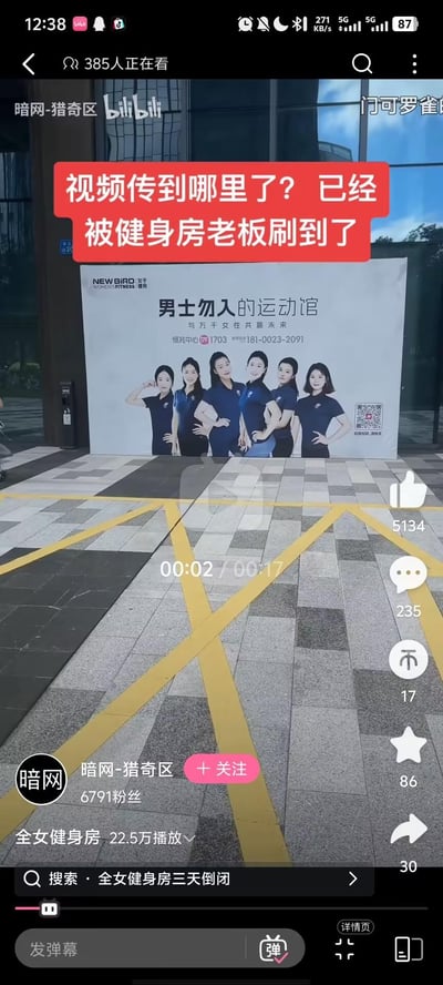 Bilibili视频截图显示一家名为“男士勿入的运动馆”的女性专属健身房外景，招牌上有六位身着蓝色制服的女性工作人员合影，下方标注联系电话。画面上方红色文字叠加“视频传到哪里了？已经被健身房老板刷到了”，暗示视频内容引发健身房经营者关注。视频来自账号“暗网-猎奇区”，界面显示385人正在观看，累计5134点赞、235评论、17分享，播放量达22.5万次。