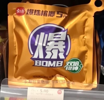 这是一张在线下零售货架拍摄的宾之郎爆珠槟榔商品实拍图，商品包装以金色为主色调，顶部左侧印有红色的“宾之郎”品牌标识，旁边标注“爆珠槟榔 5元”；包装中央是紫色圆形边框包裹的白色醒目大字“爆”，下方搭配英文“BOMB”，右侧有绿色圆形标识，内写“双倍提神”；包装底部印有生产企业“湖南宾之郎实业集团有限公司”以及“长期过量咀嚼 有害口腔健康”的健康警示语。包装下方的货架价签显示该商品售价为5.00元，整体场景是在便利店或超市的零食售卖区域。