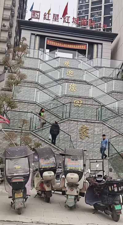 这是一张拍摄于红星·红城国际三期小区入口的照片，入口处的楼梯玻璃护栏上依次印有「欢」「迎」「您」「回」「家」的金色大字，由于楼梯的台阶式结构，从下方视角看，这组标语被楼梯分割，形成了有趣的视觉错位效果，仿佛每个字都分布在不同的台阶层面。画面下方停放着多辆带雨棚的雅迪电动车，还有几位行人正在楼梯上行走，入口上方的电子屏滚动着「乐恒物业全体员工祝红城国际业主新年快乐」的祝福语，建筑顶部清晰标注着小区名称。
这张图的趣味点在于视觉错位带来的奇妙观感，原本正常的标语因楼梯结构显得格外有意思。