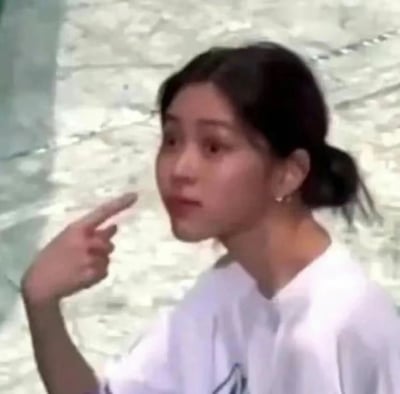 图片中是一位年轻女性，她留着黑色的头发并扎成了一个丸子头，耳朵上戴着小巧的耳环。她穿着一件白色的上衣，正用右手食指指向自己的左侧方向。她的眼睛睁大，嘴巴微张，脸上带着一丝疑惑和惊讶的表情，似乎看到了什么让她感到意外的东西。背景是浅色的大理石纹理墙面，整体画面给人一种日常抓拍的感觉。