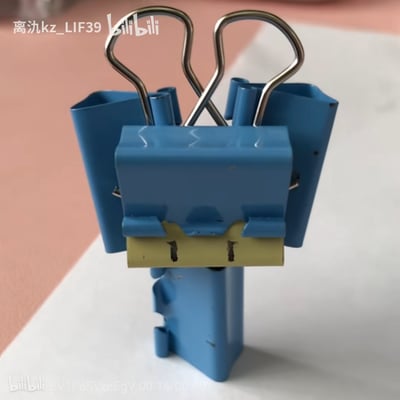 这是一张用多个蓝色长尾夹手工拼接而成的创意造型图，将长尾夹的金属夹臂作为机器人的“天线”，夹身部分组合出机器人的躯干和类似手臂的结构，把办公常用的长尾夹改造成充满趣味的机器人形象，展现了日常文具的创意玩法。