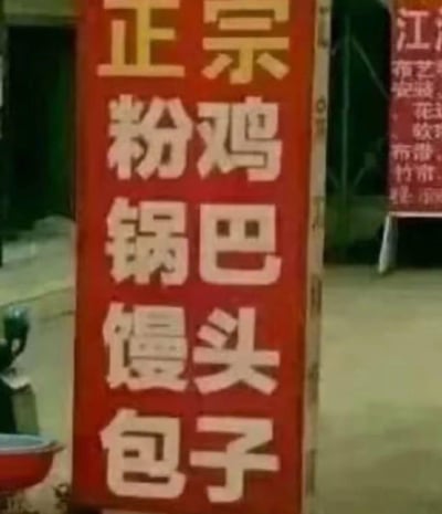 图片展示了一个街头场景中的红色立式招牌，招牌上以醒目的白色大字纵向排列着“正宗粉鸡锅巴馒头包子”，字体较大且清晰。背景中隐约可见另一块红色招牌的部分文字，但焦点集中在前景的这块主要招牌上，整体给人一种接地气的地方小吃摊氛围。