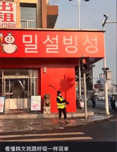 一张街头照片，展示了一家红色店面，招牌上有类似蜜雪冰城的卡通雪人形象和看似韩文的文字"밀설빙성"。一名穿着黄色安全背心的环卫工人正在店前清扫。图片底部配有文字"看懂韩文就跟呼吸一样简单"，形成幽默效果。

实际上招牌上的韩文是无意义的组合，并非真正的韩文，这种设计制造了假装懂韩文的搞笑效果。