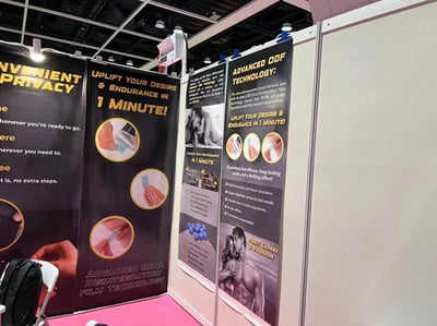 图片展示了一个贸易展会的展位，展位上有多块黑色和黄色相间的宣传横幅，主要推广一款声称能“提升欲望与耐力”的产品。横幅上有醒目的黄色大字标语，如“UPLIFT YOUR DESIRE & ENDURANCE IN 1 MINUTE!”和“ADVANCED ODF TECHNOLOGY”，并配有产品使用步骤的图示（如手部操作产品的圆形图片）、黑白情侣照片以及产品功效说明（如“快速起效、持久效果、无副作用”等）。展位前方地面为粉色，左侧有一把白色椅子和一个黑色背包。