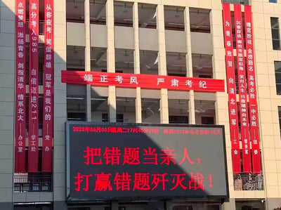 这是2023年6月6日（高考前一天）某第一中学的教学楼外实景，楼体悬挂多条红色励志横幅，内容涵盖冲刺清北、985/211的目标激励，从容应考的心态引导，还有强调考风考纪的横幅；下方电子屏显示精确到秒的高考倒计时，标注距离2023年高考仅剩0天，同时打出「把错题当亲人，打赢错题歼灭战！」的趣味励志标语，整体氛围紧张且充满斗志，是国内高中典型的考前动员场景，无谐音梗或双关内容。
