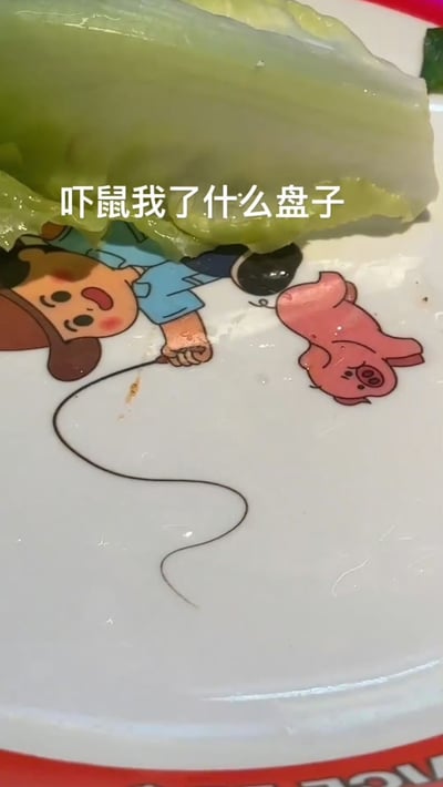 这是一张恶搞日常餐盘趣图。图片中有一张生菜叶盖在带有蜡笔小新角色图案的盘子上，而盘面上还存在一根像头发一样的异物，组合起来产生了强烈的视觉错位效果。由于生菜刚好盖住小新头部位置，看起来像小新披着绿发，而长发状异物又和小新的手或表情产生某种意义的不协调。图片配文字“吓鼠我了什么盘子”，用夸张网络热词表达惊吓和无语，整体气氛幽默、整蛊、有点恶趣味。没有显著的谐音梗、暗示，但有视觉错觉带来的错位幽默。