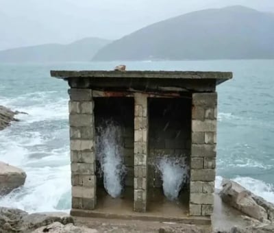 一张海边风景照，画面中央是一座简陋的石砌小屋，小屋有两个并排的洞口，海水正从两个洞口向外喷涌，形成两道白色的水柱。石屋坐落在海边的岩石上，周围是波涛汹涌的蓝色海水，远处可以看到朦胧的山脉轮廓，天空呈现出灰蒙蒙的阴天景象。整个场景给人一种奇特而壮观的感觉，仿佛这座石屋是连接海洋的神秘通道。