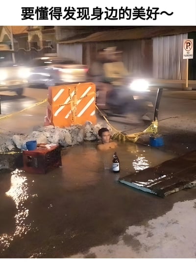 这张梗图展示了一名男子坐在马路的积水坑中，旁边放着一瓶啤酒，周围有施工路障和警示带。图片上方配有文字'要懂得发现身边的美好~'，通过幽默的方式表达了在逆境中寻找乐趣的乐观态度。