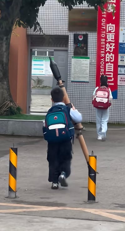这是一张实拍的上学时段校园入口照片，画面中一名背着蓝绿拼色书包的男小学生背对着镜头走在入校道路上，他的右肩扛着一个很长、前端为黑色锥形的类似火箭模型的长杆大件物品，造型格外引人注目，和普通上学的场景形成趣味反差；画面前方还有一名背着粉红色书包的女生正走向教学楼入口，道路两侧有黄黑相间的警示路桩，教学楼墙面铺有小方格瓷砖，还悬挂着红色的中英文励志标语，整体场景充满真实的生活趣味。