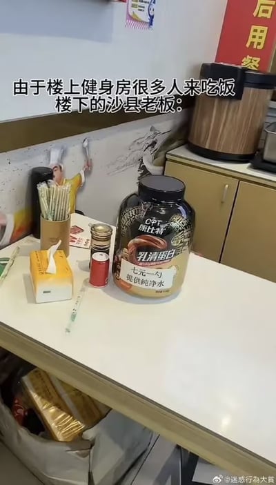 沙县小吃店内的白色柜台上，摆放着一罐康比特(CPT)乳清蛋白粉，罐子上贴着手写纸条“七元一勺 提供纯净水”。柜台周围有吸管筒、纸巾盒、调味料罐和一个注射器（可能用于取水），背景墙贴着餐饮相关海报，右侧有金色橱柜和咖啡机。图片上方文字“由于楼上健身房很多人来吃饭 楼下的沙县老板：”揭示了老板为吸引健身人群推出的特色服务，展现了商家灵活应变的幽默智慧。