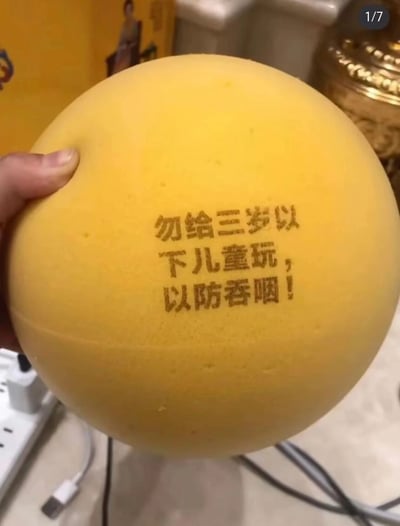 一只手握着一个黄色的球体，球体上印有警告文字："勿给三岁以下儿童玩，以防吞咽！"。图片右上角有"1/7"的标识，表明这可能是系列图片中的第一张。背景中可以看到一些模糊的家居物品，包括一个金色的装饰罐和黄色的盒子。