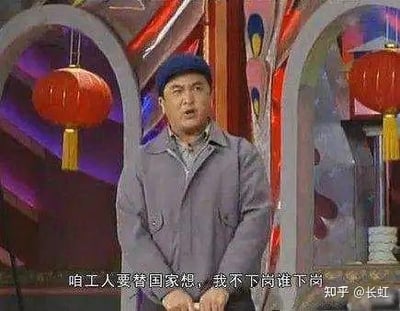 图片为春晚小品截图，一名身穿灰色夹克、头戴蓝色帽子的男性站在装饰有红色灯笼和彩色背景的舞台上，表情严肃，正在说出台词：'咱工人要替国家想，我不下岗谁下岗'。该台词反映了特定历史时期工人面对下岗的无奈与集体意识。